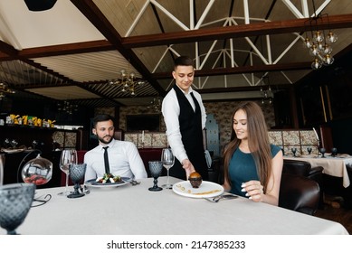 A Young Waiter In A Stylish Apron Serves A Table With A Beautiful Couple In A Refined Restaurant. Exquisite Delicacies Of Haute Cuisine Close-up.