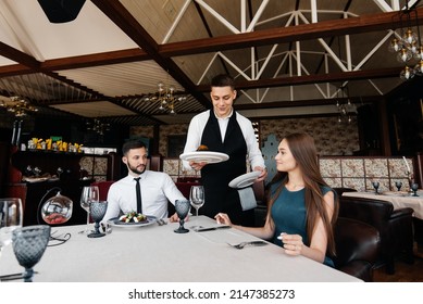 A Young Waiter In A Stylish Apron Serves A Table With A Beautiful Couple In A Refined Restaurant. Exquisite Delicacies Of Haute Cuisine Close-up.