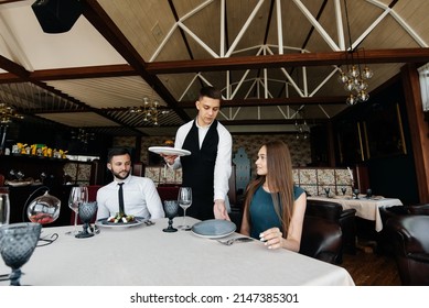 A Young Waiter In A Stylish Apron Serves A Table With A Beautiful Couple In A Refined Restaurant. Exquisite Delicacies Of Haute Cuisine Close-up.