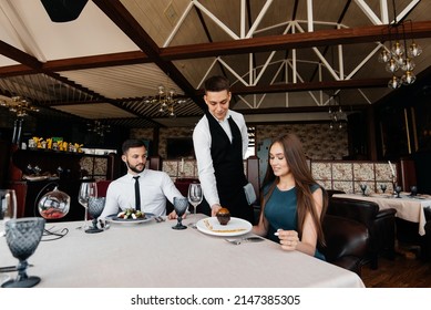 A Young Waiter In A Stylish Apron Serves A Table With A Beautiful Couple In A Refined Restaurant. Exquisite Delicacies Of Haute Cuisine Close-up.