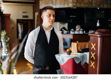 A young waiter in a stylish uniform stands with an exquisite dish on a tray near the table in a beautiful restaurant close-up. Restaurant activity, of the highest level.