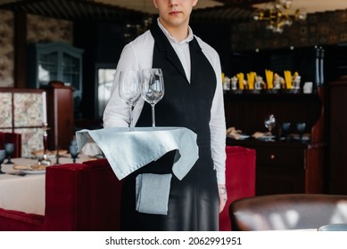 A young waiter in a stylish uniform stands with glasses on a tray near the table in a beautiful gourmet restaurant close-up. Restaurant activity, of the highest level.