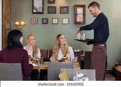 Young Waiter With Tray Of Glasses While Female Customers Having Meal In Restaurant