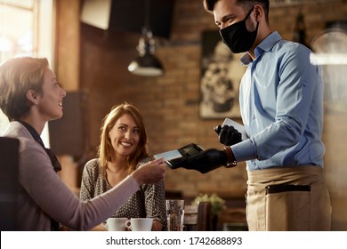 Young waiter wearing protective face mask while his guests are making contactless payment with credit card in a cafe. 