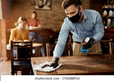 Young waiter wearing protective face mask while cleaning tables while working in a cafe. 