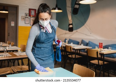 Young Waitress With Apron, Face Mask And Gloves Cleaning Tables Restaurant With Sanitizer And Cloth