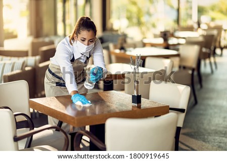 Young waitress disinfecting tables while wearing protective face mask ad gloves due to coronavirus epidemic. 