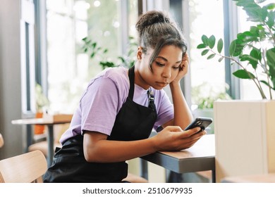 Young waitress looks stressed while checking her smartphone in a cozy cafe, showing a mix of emotions in a small business setting. Online technology concept - Powered by Shutterstock - Get 15% off with code: PIKWIZARD15