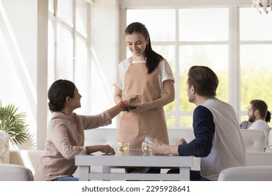 Young waitress and positive Indian female customer connecting her modern smartwatch to payment terminal equipment, client makes easy, comfort cashless wireless payment sits at table in cafe. NFC tech - Powered by Shutterstock