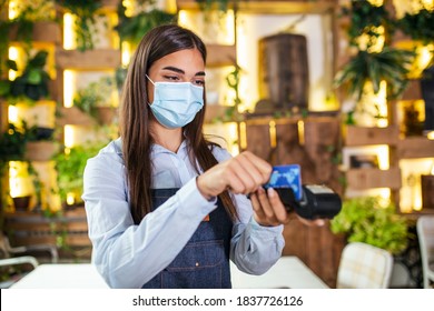 Young Waitress With Protective Face Mask Making A Credit Card Payment With Card Reader Machine. Credit Card Swipe Through Terminal In Restaurant. Waitress Hand Swiping Debit Card To Pay The Bill