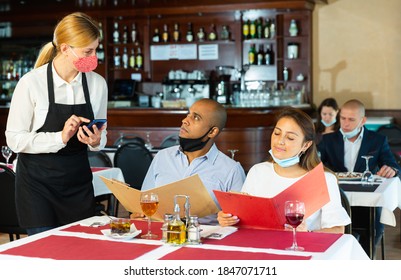 Young Waitress In Protective Face Mask Taking Order, Helping Hispanic Couple In Choosing Meals From Menu. Concept Of Reopening Of Restaurants After Quarantine