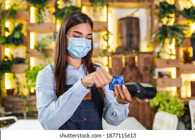 Young Waitress With Protective Face Mask Making A Credit Card Payment With Card Reader Machine. Credit Card Swipe Through Terminal In Restaurant. Waitress Hand Swiping Debit Card To Pay The Bill