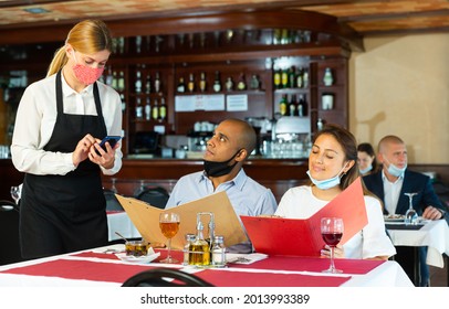 Young Waitress In Protective Face Mask Taking Order, Helping Hispanic Couple In Choosing Meals From Menu. Concept Of Reopening Of Restaurants After Quarantine