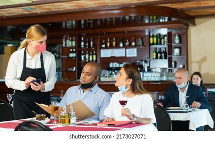 Young Waitress In Protective Face Mask Taking Order, Helping Hispanic Couple In Choosing Meals From Menu. Concept Of Reopening Of Restaurants After Quarantine