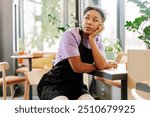 Young waitress is sitting alone at a table in a quiet coffee shop, resting her chin on her hand and looking away with a thoughtful expression