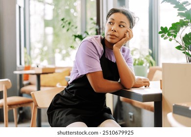 Young waitress is sitting alone at a table in a quiet coffee shop, resting her chin on her hand and looking away with a thoughtful expression - Powered by Shutterstock