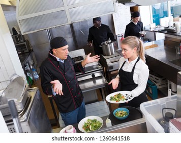 Young Waitress Standing With Cooked Dish In Restaurant Kitchen Talking With Puzzled Male Chef