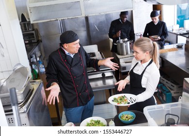 Young Waitress Standing With Cooked Dish In Restaurant Kitchen Talking With Puzzled Male Chef