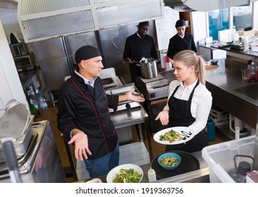 Young Waitress Standing With Cooked Dish In Restaurant Kitchen Talking With Puzzled Male Chef