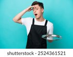 Young waitress with tray isolated on blue background doing surprise gesture while looking to the side