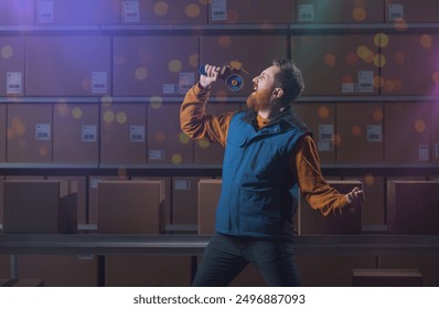 Young warehouse worker singing instead of working, he is using the hand tape dispenser as microphone