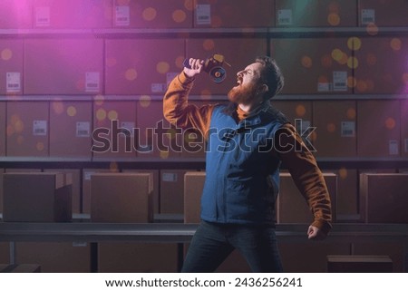 Young warehouse worker singing instead of working, he is using the hand tape dispenser as microphone