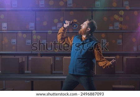Young warehouse worker singing instead of working, he is using the hand tape dispenser as microphone