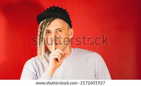 Young watchful man with blonde dreadlocks in black cap looking at camera, putting his forefinger to mouth on red background. Cautious guy showing gesture of silence with index finger at lips