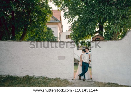 Young wedding couple outdoors near the white concrete fence, wall. Bride in wonderful wedding dress hugging groom. Countryside