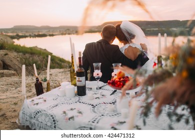 Young Wedding Couple Siiting Back To Lake At Romantic Evening With Candles On The Sand Beach