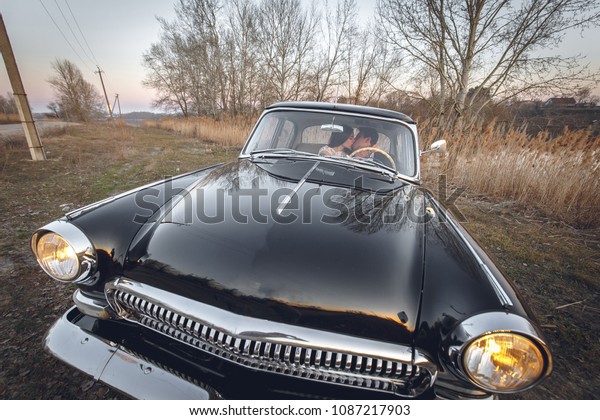 Young wedding couple sitting smiling inside retro\
car and looking at each other. just married embrace is hugging\
inside car. bride hugging groom who is driving the car. Wedding\
with retro car.