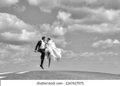 Young Wedding Happy Couple Of Sexy Girl With Brunette Hair And Pretty Face In White Bride Dress And Handsome Man In Black Groom Suit Running On Road Way On Cloudy Blue Sky Background, Copy Space