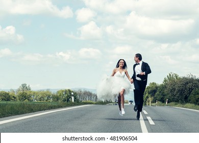 Young Wedding Happy Couple Of Sexy Girl With Brunette Hair And Pretty Face In White Bride Dress And Handsome Man In Black Groom Suit Running On Road Way On Cloudy Blue Sky Background, Copy Space