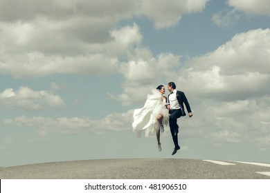 Young Wedding Happy Couple Of Sexy Girl With Brunette Hair And Pretty Face In White Bride Dress And Handsome Man In Black Groom Suit Running On Road Way On Cloudy Blue Sky Background, Copy Space