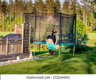 Young well trained sporty teenager performs acrobatic exercise salto - flip backwards, feet up, hands around knees. Exercise on green grass near wooden terrace fence and round trampoline, summer - Shutterstock ID 1722244351