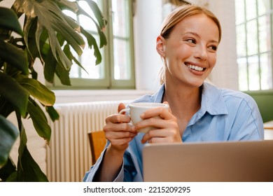 Young white businesswoman drinking coffee and using laptop while sitting by table in cafe - Powered by Shutterstock