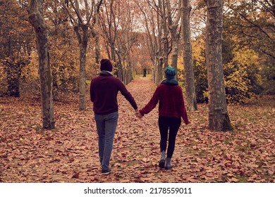 Young White Caucasian Couple Walking Backwards Holding Hands Apart Strolling Along A Ground Of Fallen Brown Leaves And A Path Of Trees In A Park In Autumn