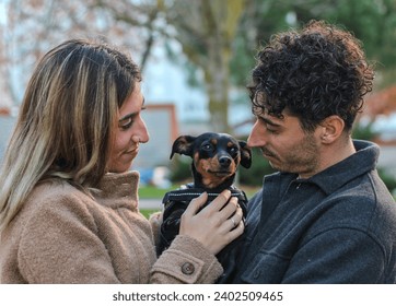 Young white couple holding a dog in a park, looking at it tenderly. Dog looking at the horizon