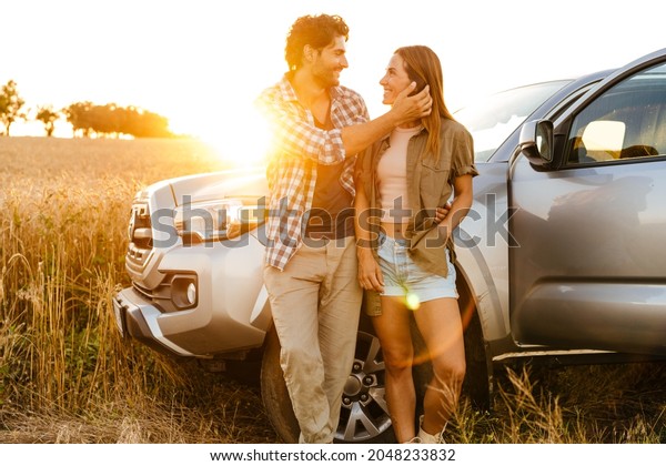 Young white couple hugging and smiling while\
standing by car on field