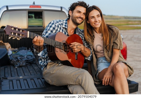 Young white couple playing guitar while sitting\
in trunk during journey\
outdoors