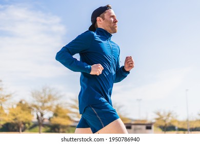 A young white european and athletic man is running in the street on a sunny day. The guy is wearing blue clothes and a cap.