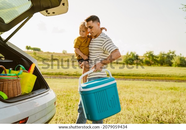 Young white father holding his son and\
cooler bag while standing by car trunk\
outdoors