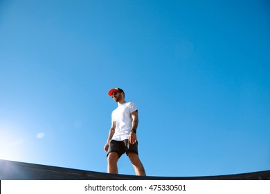 Young White Guy With Beard And Tattoos Wearing A Plain White T-shirt, Shorts, Baseball Hat And Sunglasses, Walking In A Skate Park Shot From Below Against Bright Blue Sky