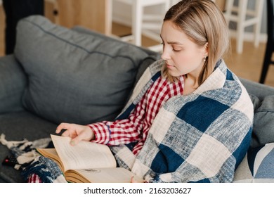 Young white happy woman wrapped in blanket reading book while sitting on sofa at home