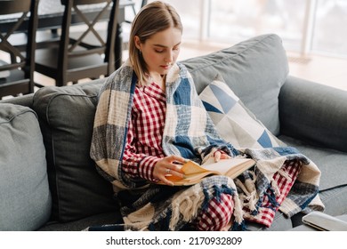 Young white happy woman wrapped in blanket reading book while sitting on sofa at home