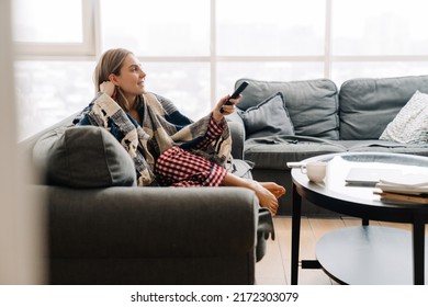 Young white happy woman wrapped in blanket holding remote control while watching television on sofa at home