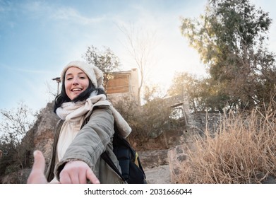 Young White Hispanic Latin Girl With Beige Cap And Scarf, Green Jacket And Backpack In Front Of A Cottage On The Edge Of A Mountain Smiling And Reaching Out Her Hand To Grab Someone, Sky In Background