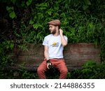 Young White Man with Beard Using a Brown Cap and Shirt Reading "Save Animals" on a Bench Surrounded by Vegetation