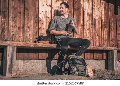 Young White Man Having A Coffee Break From Hiking In Swedish Östersund During Outdoor Advebture. Sitting On A Wooden Bench Looking Happily Sidewards.