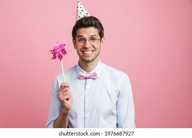 Young White Man Wearing Party Cone Posing With Flower Wind Spinner Isolated Over Pink Background